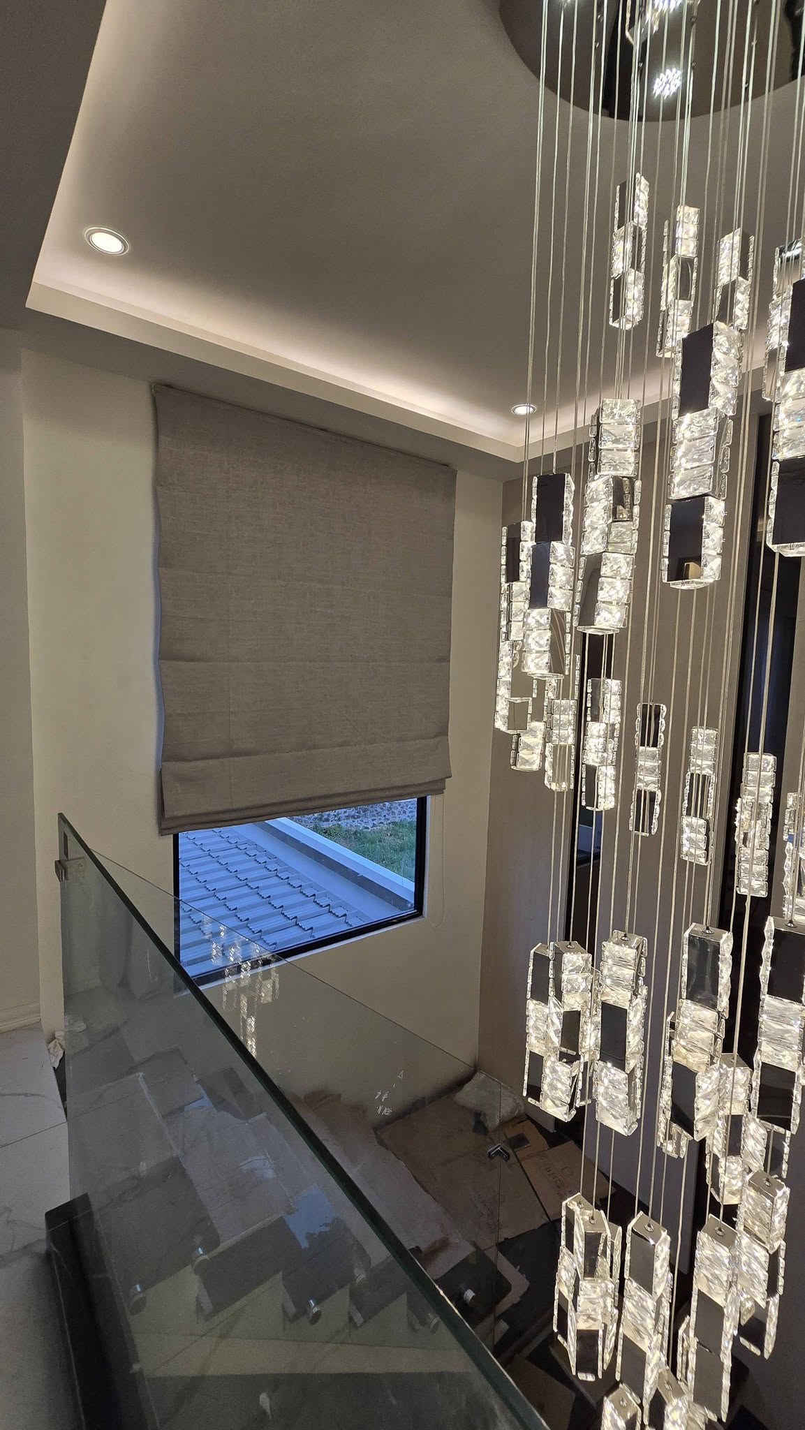 Premium linen-texture Roman blind in neutral taupe, installed beside glass staircase and luxury chandelier