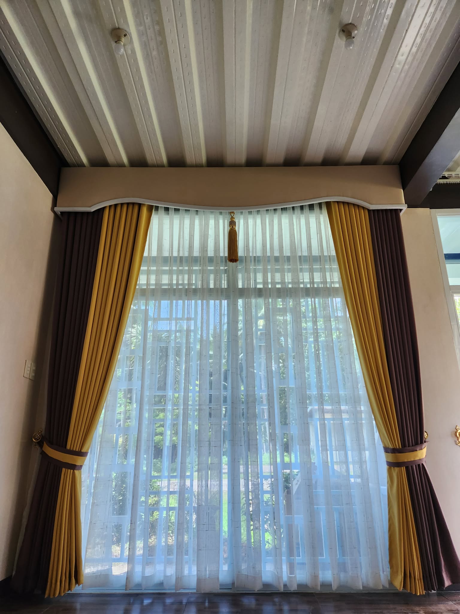 Window with two-tone brown and gold drapes, white sheer curtains, and custom curved pelmet